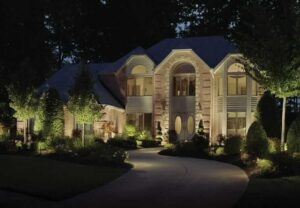 A large, two-story brick house is illuminated at night by expertly installed fixtures, part of an outdoor lighting franchise, surrounded by trees and landscaped gardens.