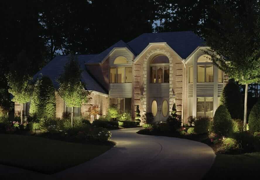 A large, two-story brick house is illuminated at night by expertly installed fixtures, part of an outdoor lighting franchise, surrounded by trees and landscaped gardens.