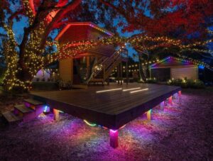 Outdoor wooden deck and treehouse illuminated with colorful string lights at night, surrounded by trees.
